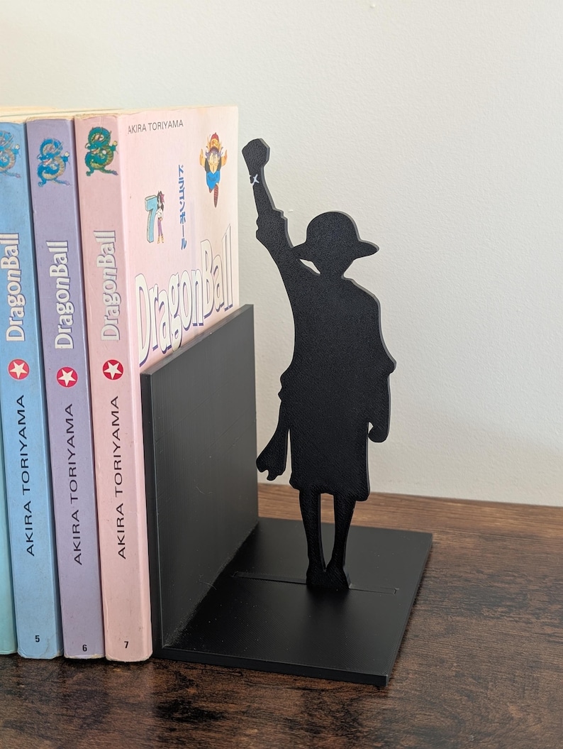 May include: A black metal bookend featuring a silhouette of a character wearing a hat with one arm raised in the air. The bookend is positioned next to three Dragon Ball manga volumes by Akira Toriyama, which have light blue, purple, and pink spines.