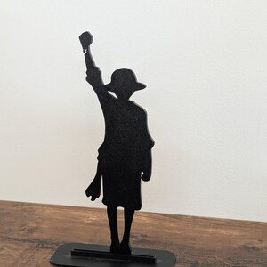 May include: A black silhouette figure of a character wearing a wide-brimmed hat, standing with one arm raised in a fist. The figure is mounted on a rectangular black base, creating a flat, two-dimensional decorative display piece.