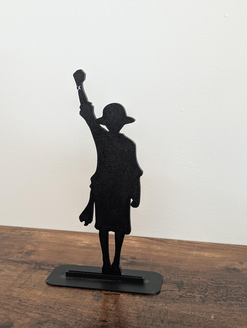May include: A black silhouette figure of a character wearing a wide-brimmed hat, standing with one arm raised in a fist. The figure is mounted on a rectangular black base, creating a flat, two-dimensional decorative display piece.