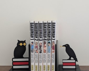May include: A set of black metal bookends featuring a silhouette of an owl wearing yellow glasses on the left and a crow holding a gold key on the right, both perched on stacks of books with red and white stripes, holding six Bakuman manga volumes.