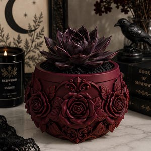 May include: A dark purple succulent plant potted in a deep red, round planter. The exterior of the planter features a relief design of sculpted roses and intricate scrollwork patterns surrounding the entire circumference of the vessel.