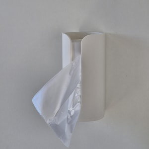 Può includere: Un distributore di sacchetti di plastica bianco, montato a parete. Il distributore è cilindrico con una parte superiore curva e una fessura per l'erogazione dei sacchetti. Un sacchetto di plastica bianco è parzialmente estratto.