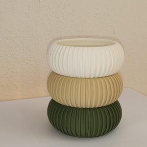 Può includere: Tre vasi in ceramica impilati di diverse dimensioni. Il vaso superiore è bianco, il vaso intermedio è beige e il vaso inferiore è verde. Tutti e tre i vasi hanno un design a coste.