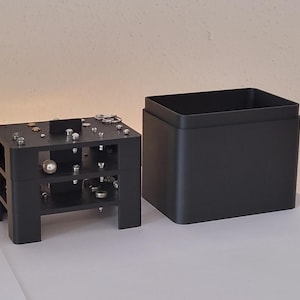 Könnte beinhalten: Eine schwarze Plastikbox mit einem abnehmbaren Deckel. Die Box ist geöffnet und der Deckel liegt daneben. Die Box hat eine Reihe kleiner Löcher und Metallteile im Inneren.