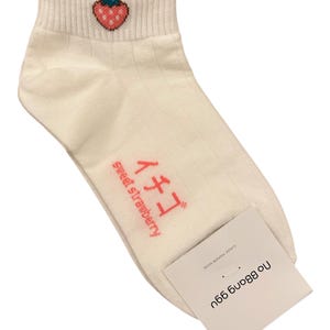 May include: Cream-colored ankle socks with a ribbed texture. The socks feature a small embroidered strawberry on the cuff and Japanese text with the words "sweet strawberry". A white tag with text is attached.