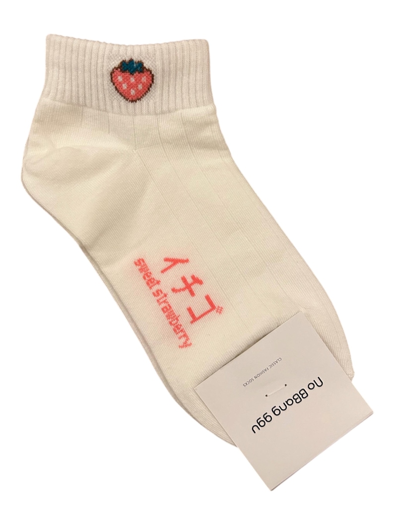 May include: Cream-colored ankle socks with a ribbed texture. The socks feature a small embroidered strawberry on the cuff and Japanese text with the words "sweet strawberry". A white tag with text is attached.