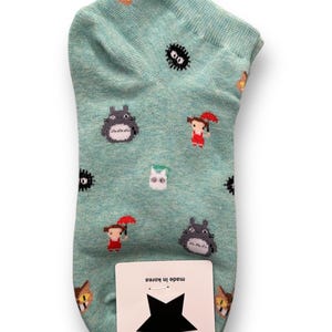 Op de afbeelding: Een lichtblauwe enkelsok met een herhaald patroon van stripfiguren. De sok heeft een grijs Totoro-achtig wezen, een meisje met een rode paraplu en andere grillige figuren. Een wit label met een zwarte ster en de tekst "Made in Korea" is bevestigd.