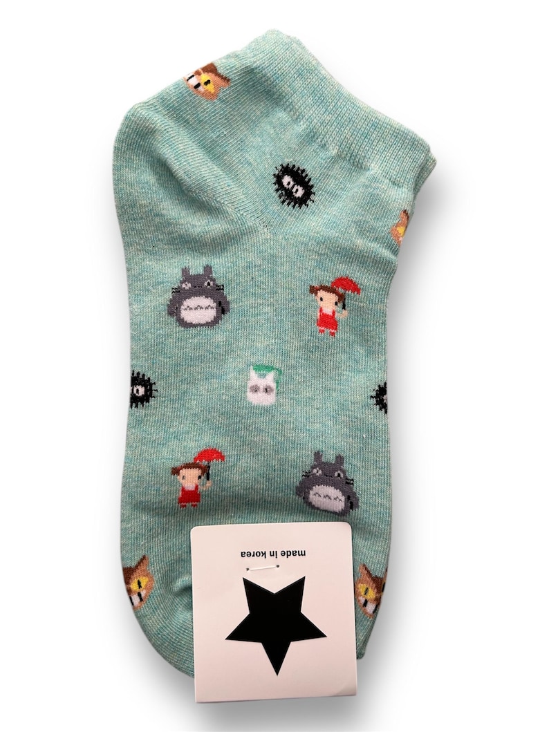 Op de afbeelding: Een lichtblauwe enkelsok met een herhaald patroon van stripfiguren. De sok heeft een grijs Totoro-achtig wezen, een meisje met een rode paraplu en andere grillige figuren. Een wit label met een zwarte ster en de tekst "Made in Korea" is bevestigd.