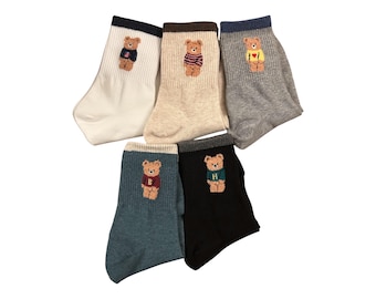 May include: Five pairs of socks in various colors, each featuring an embroidered teddy bear design. The socks are white, beige, gray, teal, and black, with the bear wearing different colored sweaters. The socks are made of a soft material.