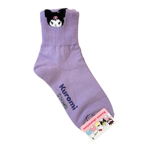 May include: Purple ankle socks with a black and white Kuromi character embroidered on the top. The socks have the text "Kuromi &copy;'21 SANRIO" printed on the side. The socks have a pink tag with the text "Sanrio Characters" and a group of Sanrio characters.