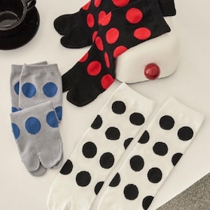 Può includere: Una collezione di calzini tabi in vari colori e fantasie. I calzini includono nero con pois rossi, bianco con pois neri e grigio con pois blu. I calzini sono esposti su una superficie bianca.