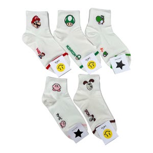 May include: Five pairs of white ankle socks featuring embroidered character graphics including Mario, a mushroom, Yoshi, Kirby, and Gromit. Each sock displays the character name in text and includes a small attached tag with a smiley face or star.