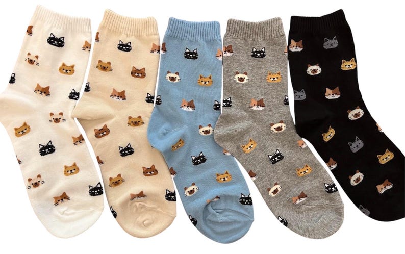 Puede incluir: Cinco pares de calcetines en varios colores: blanco roto, beige, azul claro, gris y negro. Cada calcet&iacute;n presenta un patr&oacute;n repetido de caras de gato en diferentes colores. Los gatos tienen pelaje negro, marr&oacute;n y canela.