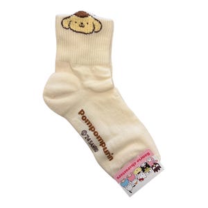 May include: Cream-colored ankle socks with a brown Pompompurin character embroidered on the top. The socks have the text "Pompompurin" and "&copy;'24 SANRIO" printed on the side. The socks have a pink tag with the text "Sanrio Characters" and images of various Sanrio characters.