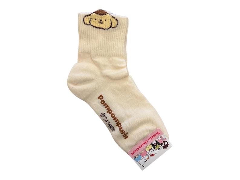 May include: Cream-colored ankle socks with a brown Pompompurin character embroidered on the top. The socks have the text "Pompompurin" and "&copy;'24 SANRIO" printed on the side. The socks have a pink tag with the text "Sanrio Characters" and images of various Sanrio characters.