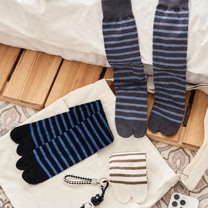 May include: A collection of split-toe socks featuring various striped patterns. One pair is dark gray with light blue stripes, another is black with bright blue stripes, and a third is white with brown stripes, accompanied by a small bear keychain.