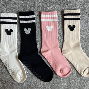 May include: Four pairs of crew-length socks featuring ribbed cuffs with two horizontal stripes. Each sock displays a small mouse head silhouette icon. The socks come in white, black, pink, and beige, with small circular grip dots on the soles.