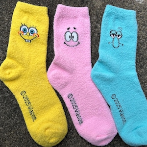 May include: Three colorful socks featuring cartoon characters. A yellow sock with SpongeBob SquarePants, a pink sock with Patrick Star, and a blue sock with Squidward Tentacles. Each sock has the text "© 2025 Viacom."