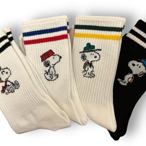 May include: Four pairs of white and black striped crew socks with different embroidered Peanuts characters. The characters are wearing different hats and outfits.