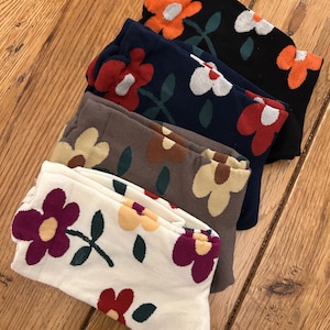 Puede incluir: Un conjunto de cinco pares de calcetines con un patrón floral. Los calcetines son de varios colores, incluyendo blanco, marrón, azul marino, negro y naranja. Cada calcetín presenta un gran diseño floral con un centro blanco.