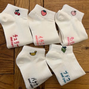 May include: Five pairs of white ankle socks with embroidered fruit designs: cherry, strawberry, peach, grape, and banana. Each sock has Japanese text and English translations. The socks are made of a soft, ribbed material.