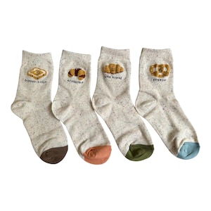 Könnte beinhalten: Vier Paar cremefarbene Socken mit bunten Sprenkeln. Jede Socke zeigt ein kleines gesticktes Lebensmittelmotiv und den entsprechenden Text: butter toast, croissant, salt bread und pretzel. Die Fersen sind braun, orange, grün und blau abgesetzt.