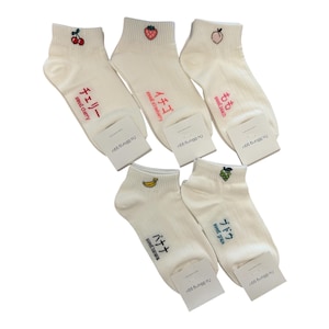 May include: A collection of white ankle socks with embroidered fruit designs, including cherries, strawberries, a peach, a banana, and grapes. Each sock features Japanese text and the English translation of the fruit name.