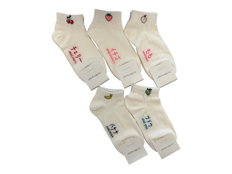 May include: A collection of white ankle socks with embroidered fruit designs, including cherries, strawberries, a peach, a banana, and grapes. Each sock features Japanese text and the English translation of the fruit name.