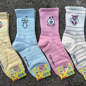 Könnte beinhalten: Vier Paar Crew-Socken mit Figuren aus der Zeichentrickserie. Die Socken sind gelb, hellblau, rosa und weiß, jede mit dem Gesicht einer Figur auf dem Bündchen gestickt. Jede Socke hat horizontale Streifen und ein kleines Etikett.