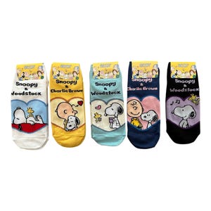 May include: Five pairs of colourful socks featuring Snoopy, Charlie Brown, and Woodstock characters. Each pair has a different design, with Snoopy in various poses and heart shapes. The socks have yellow tags with the Snoopy logo.