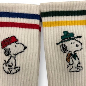May include: Two pairs of white crew socks with colorful stripes. One pair features a cartoon Snoopy wearing a red hat, the other pair features Snoopy wearing a green hat.