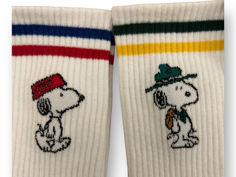 May include: Two pairs of white crew socks with colorful stripes. One pair features a cartoon Snoopy wearing a red hat, the other pair features Snoopy wearing a green hat.