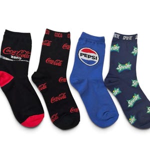 Pode incluir: Um conjunto de quatro meias com logótipos de marcas. Uma meia preta tem o logótipo Coca-Cola Zero com calcanhar e biqueira vermelhos. Uma meia preta tem o logótipo Coca-Cola vermelho repetido. Uma meia azul tem o logótipo circular Pepsi. Uma meia azul escura tem o logótipo Sprite repetido.