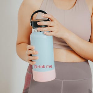 May include: A light blue reusable water bottle with a black lid and a pink silicone sleeve. The bottle has the text "Drink me." printed on it.