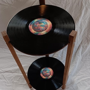 May include: A two-tiered stand made of wood with two black vinyl records as the shelves. The records have colorful, abstract designs in the center.
