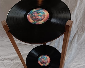 Pode incluir: Um suporte de dois níveis feito de madeira com dois discos de vinil pretos como prateleiras. Os discos têm desenhos abstratos coloridos no centro.