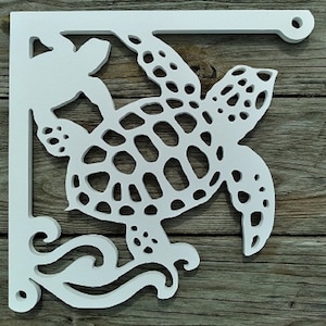 Puede incluir: Estante esquinero de madera blanca con diseño de tortuga marina. El estante tiene un patrón de olas decorativas y un agujero circular para colgar. La tortuga marina tiene un caparazón detallado con múltiples agujeros recortados.