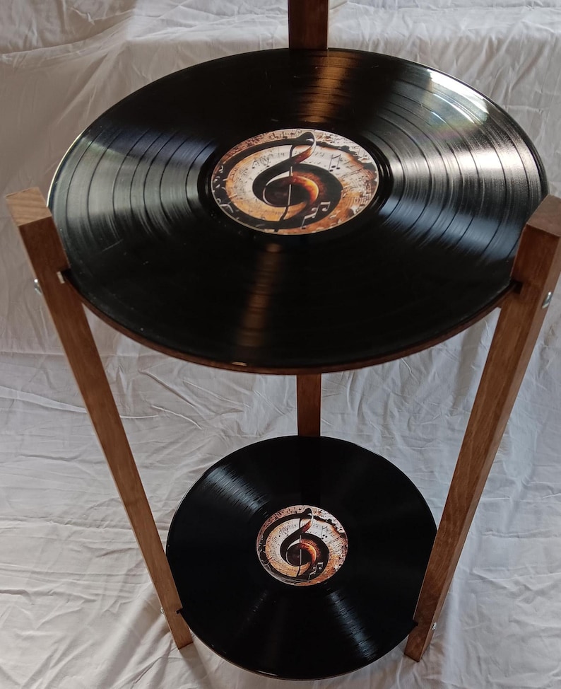 May include: A two-tiered side table made from repurposed vinyl records. The table features a wooden frame and two circular shelves, each topped with a black vinyl record. The top shelf has a decorative design of a treble clef and musical notes.
