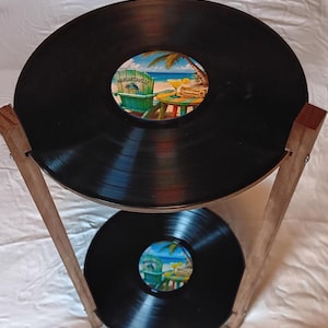 May include: A two-tiered stand constructed from wooden supports holding two black vinyl records. Each record features a central label depicting a beach scene with a green chair, a palm tree, and a cocktail, with the text Margaritaville visible.