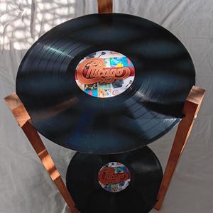 May include: A two-tiered stand constructed from brown wooden legs featuring two black vinyl records as shelves. Each record has a central label with a collage of images and the word Chicago printed in a stylized orange and white script font.