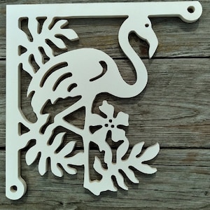 Op de afbeelding: Een witte, decoratieve hoekbeugel met een flamingo-ontwerp met tropische bladeren en bloemen. De beugel is gemaakt van hout en heeft een uitgesneden ontwerp. Ontworpen om aan een muur te worden bevestigd.