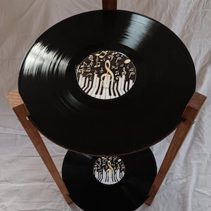 May include: A two-tiered stand made of wood with two black vinyl records as the shelves. The top record has a sticker with a treble clef and piano keys, surrounded by musical notes.