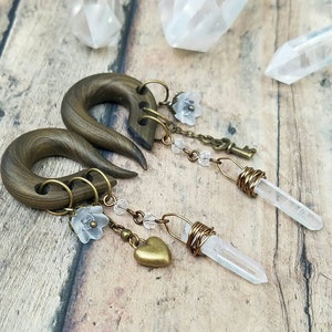 May include: A pair of brown wooden ear gauges with dangling charms. The charms include clear quartz crystals, small white flowers, a heart charm, and a small key charm.