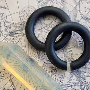 May include: Two black wooden hoop earrings, one larger than the other, are displayed on a vintage map background. The earrings are both matte black and have a smooth, rounded surface.