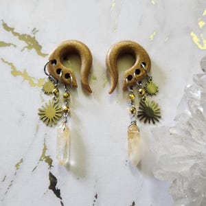 May include: A pair of golden-colored, curved ear gauges with dangling charms. Each gauge features a sunburst charm, gold beads, and a clear, elongated crystal. The gauges are displayed against a white and gold marbled background.