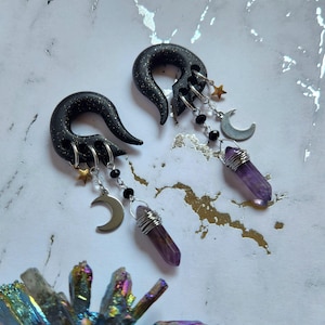 Puede incluir: Un par de expansores de oreja negros y plateados con colgantes, incluyendo medias lunas, estrellas y puntas de cristal morado.
