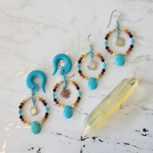 May include: A pair of turquoise dangle earrings with a sun charm and colorful beads. The earrings are made with turquoise, orange, yellow, and black beads. The earrings are hanging from a turquoise hook.
