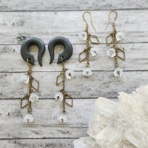 May include: A pair of black wood ear gauges with dangling chains and white flower charms. The chains are adorned with small diamond-shaped charms.