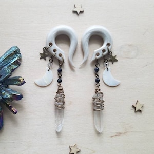 May include: A pair of white wooden ear gauges with dangling charms. The charms include a crescent moon, stars, and a clear quartz crystal wrapped in wire. The gauges are hanging from a wooden surface.