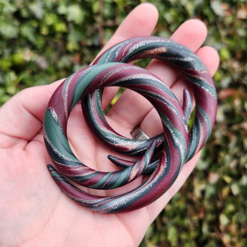 Peut inclure: Deux jauges d'oreille &agrave; motifs tourbillonn&eacute;s rouge, vert et argent. Les jauges sont en argile polym&egrave;re et ont une finition brillante.